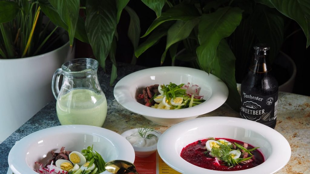 A sophisticated dining table setup featuring beetroot soup, salads, a bottle of drink, and a pitcher amidst lush greenery.