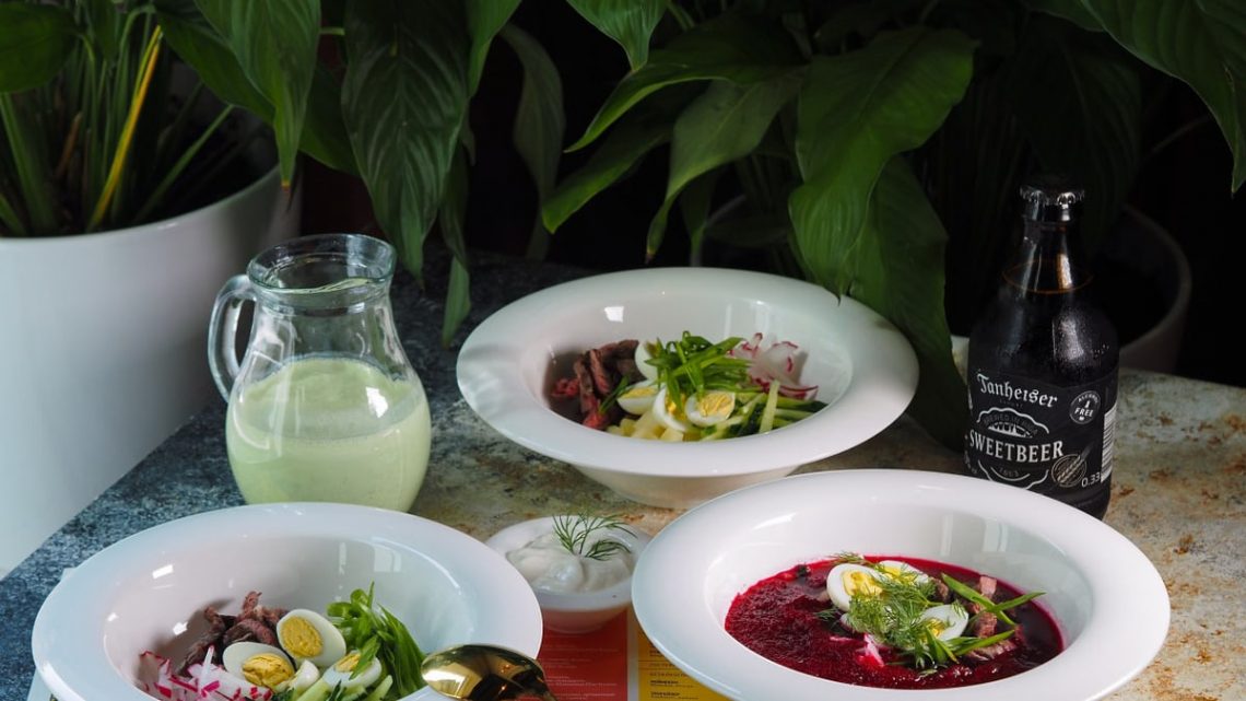 A sophisticated dining table setup featuring beetroot soup, salads, a bottle of drink, and a pitcher amidst lush greenery.