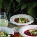 A sophisticated dining table setup featuring beetroot soup, salads, a bottle of drink, and a pitcher amidst lush greenery.